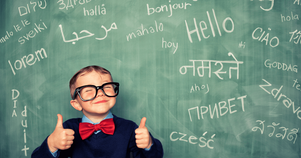 A young boy wearing glasses and a red bow tie stands in front of a chalkboard covered in greetings from different languages, smiling and giving two thumbs up—celebrating the joys of dual language learning.