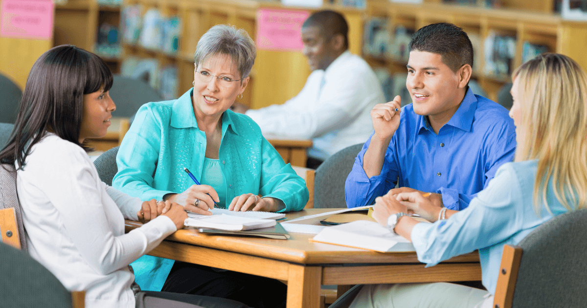 Four adults sit around a table in a library, engaged in lively advocacy and discussion. They have notebooks and papers in front of them, while others are seen working in the background.