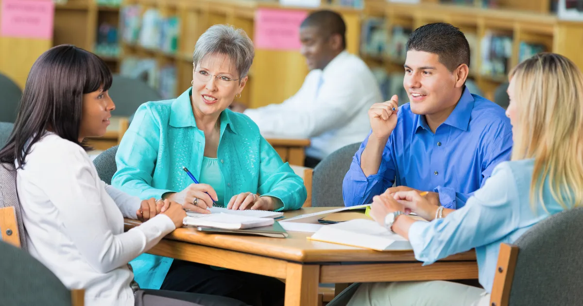 Four adults sit around a table in a library, engaged in lively advocacy and discussion. They have notebooks and papers in front of them, while others are seen working in the background.