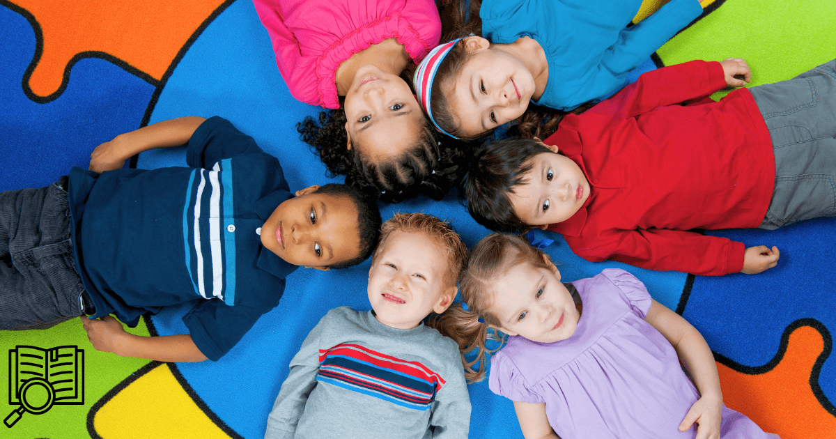 Six young children lay in a circle on a colorful puzzle-piece mat, heads together and feet outward, smiling up at the camera. An open book and magnifying glass graphic in the corner hints at their budding interest in research and discovery.