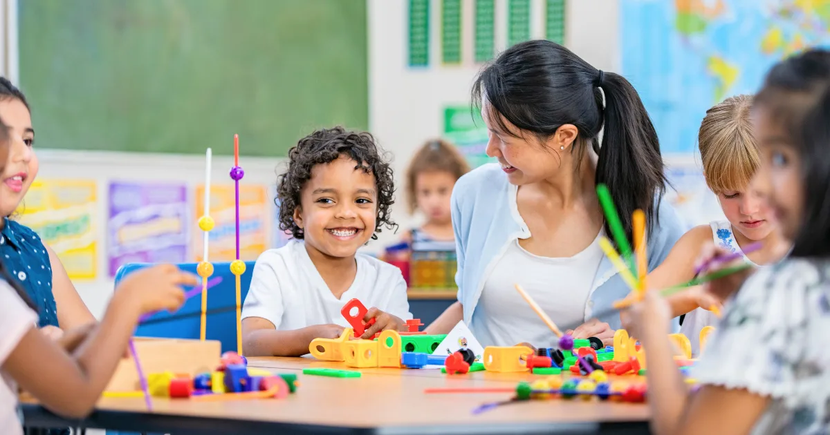 A teacher and several young children sit at a classroom table, smiling and playing with colorful educational toys and building blocks—an engaging scene of early childhood education. The background shows posters and a chalkboard.