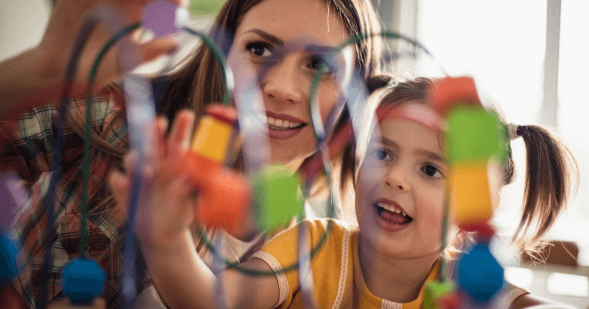 A woman and a young girl with pigtails play with a colorful bead maze toy together, smiling and focused, in a bright indoor setting—an engaging moment that could be observed during a program evaluation.