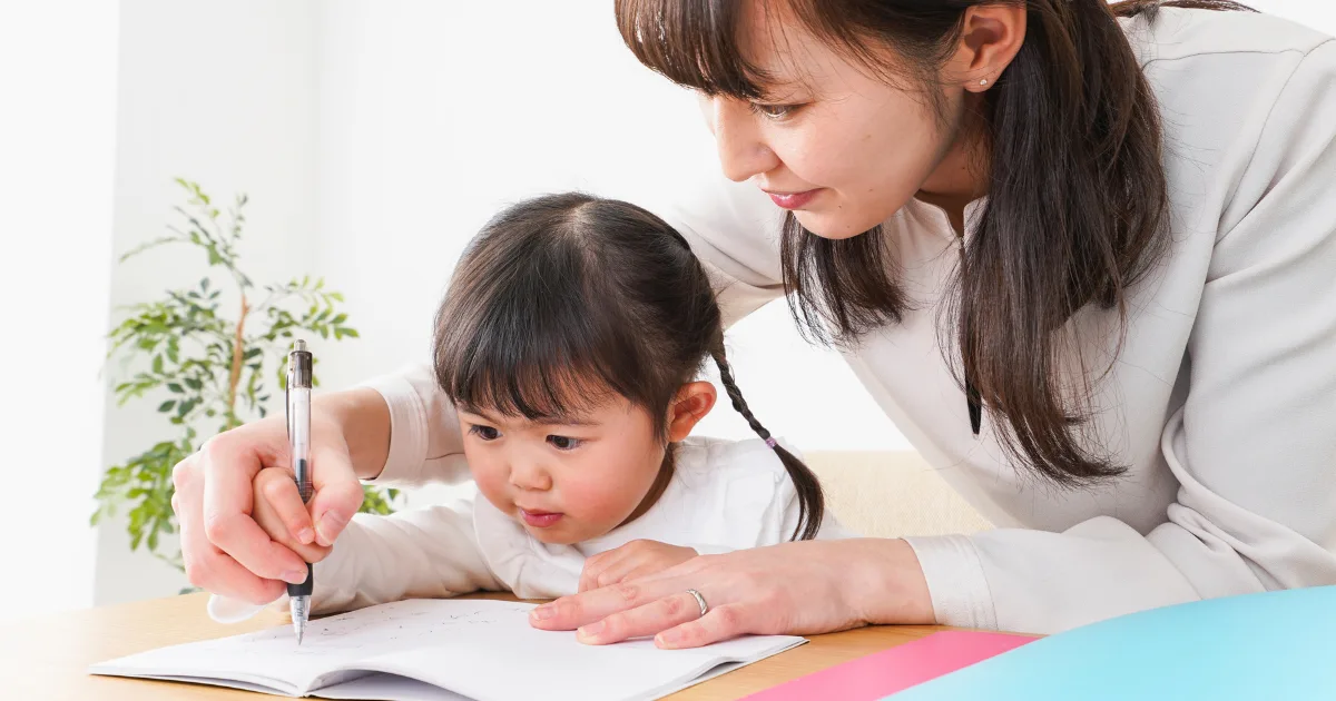 A woman helps a young girl write in a notebook at a table, reflecting their program philosophy. The woman guides the girl's hand with a pen as both focus on the page. A green plant is in the background.