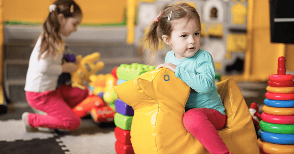 Two young children play in a colorful indoor play area at one of the early learning centers near me. One rides a yellow rocking duck, while the other plays with toys on the floor. Bright rings and blocks add to the fun atmosphere.
