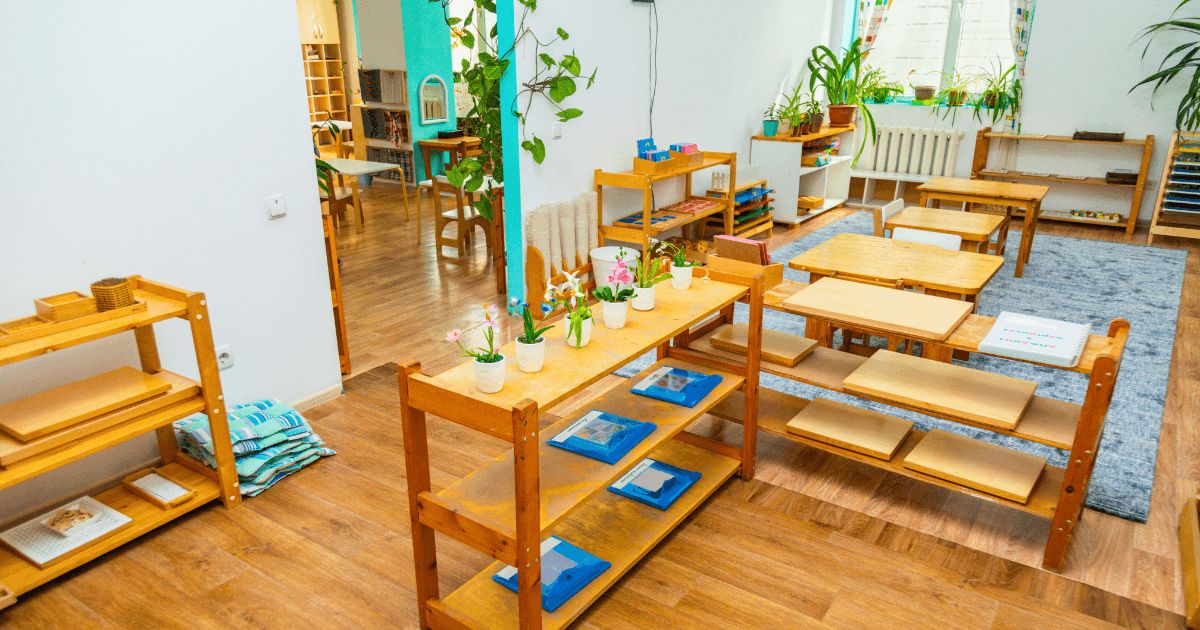 A bright, organized Montessori classroom with eco-friendly wooden shelves, educational materials, plants, child-sized tables and chairs, and sunlight streaming through windows with colorful curtains.