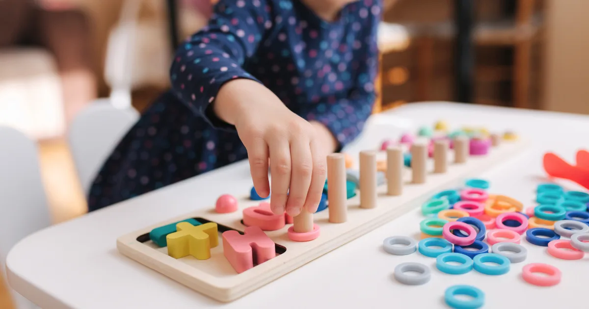 A young child in a blue, polka-dotted dress plays with a colorful wooden sorting toy at a white table, enjoying educational games by placing rings and shapes onto vertical pegs.
