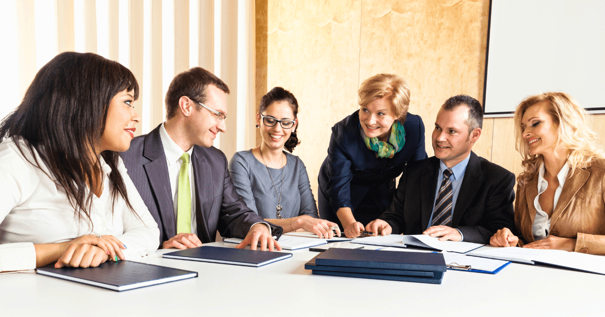 Six business professionals, three men and three women, sit around a table with documents and folders, smiling and engaged in a lively staff meeting in a bright office setting.