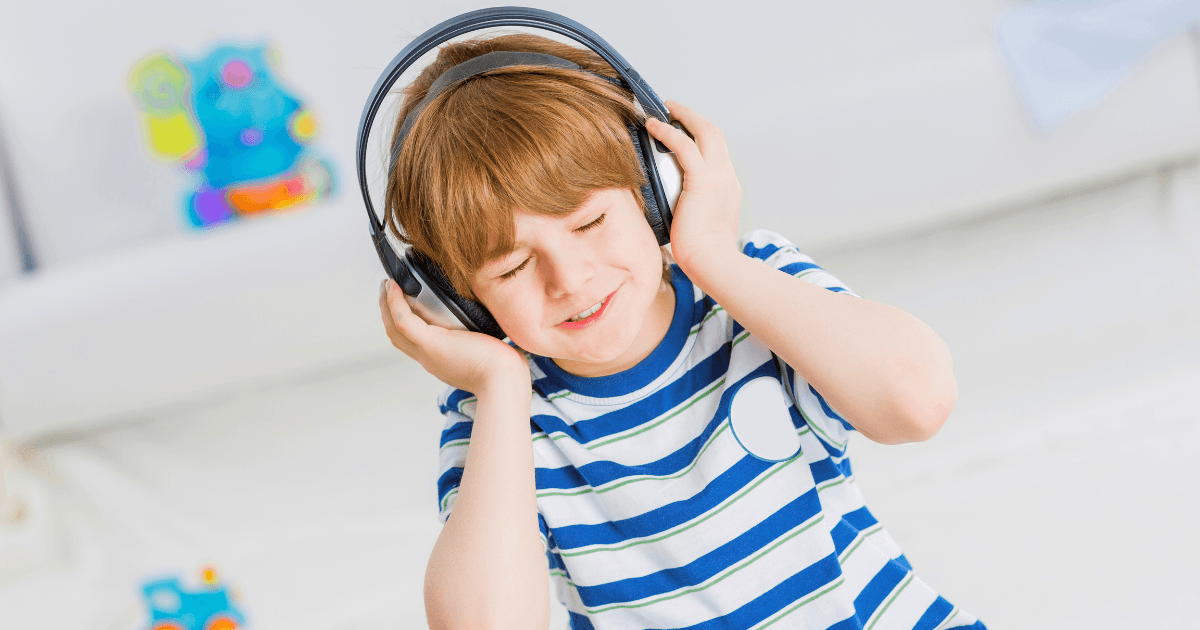A young child with light brown hair wearing a striped shirt listens to music on large black headphones, smiling with eyes closed while sitting indoors—an uplifting moment of emotional first aid. Colorful toys are blurred in the background.