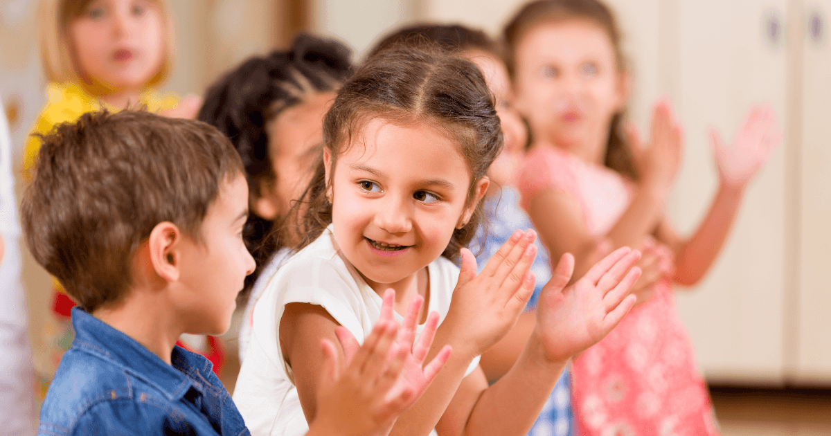 A group of young children are standing in a classroom, smiling and clapping their hands together, showcasing emotional intelligence in preschoolers. The main focus is on a girl in the center looking at a boy beside her, with others blurred in the background.