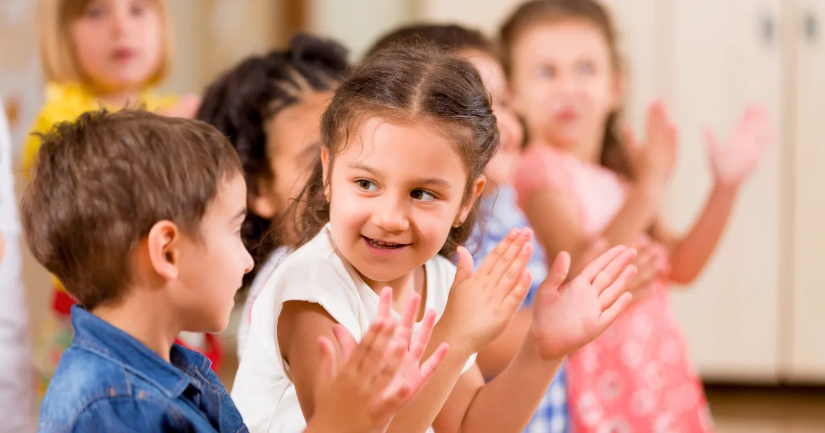 A group of young children are standing in a classroom, smiling and clapping their hands together, showcasing emotional intelligence in preschoolers. The main focus is on a girl in the center looking at a boy beside her, with others blurred in the background.