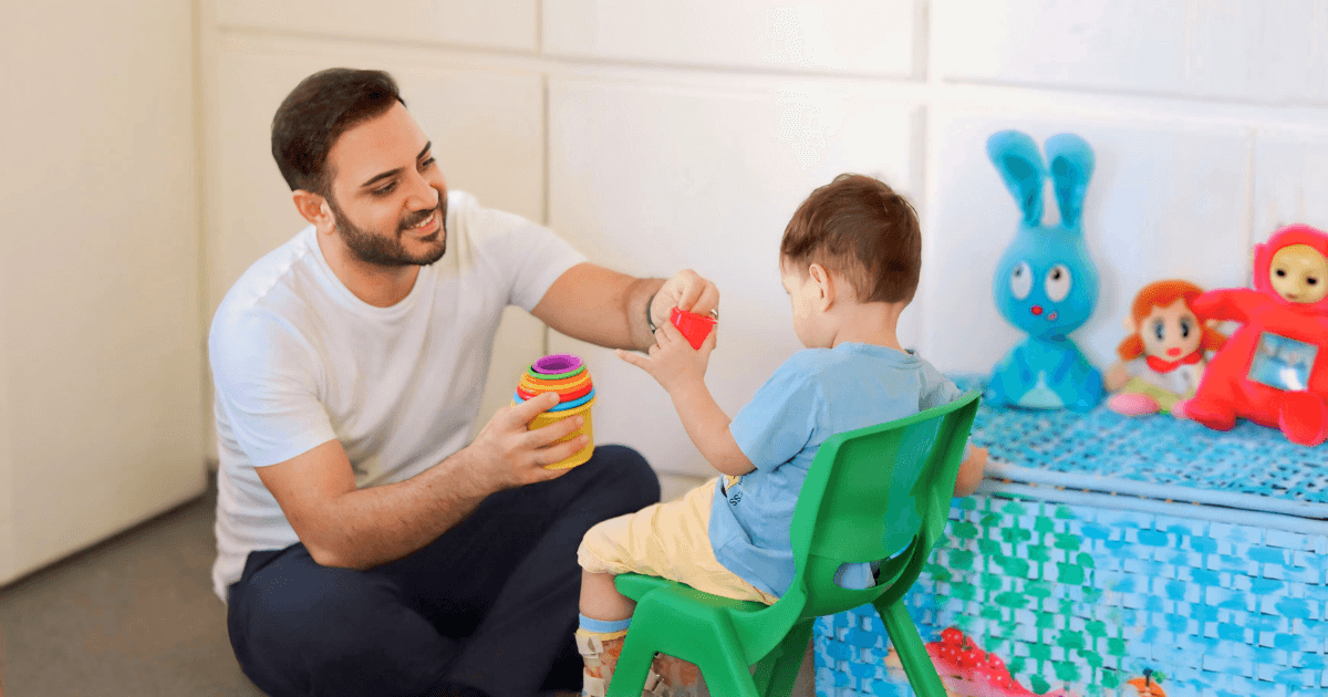 A man smiles while engaging fathers' role, playing with a young child seated on a green chair. They are stacking colorful rings, with plush toys and a blue storage basket in the background.