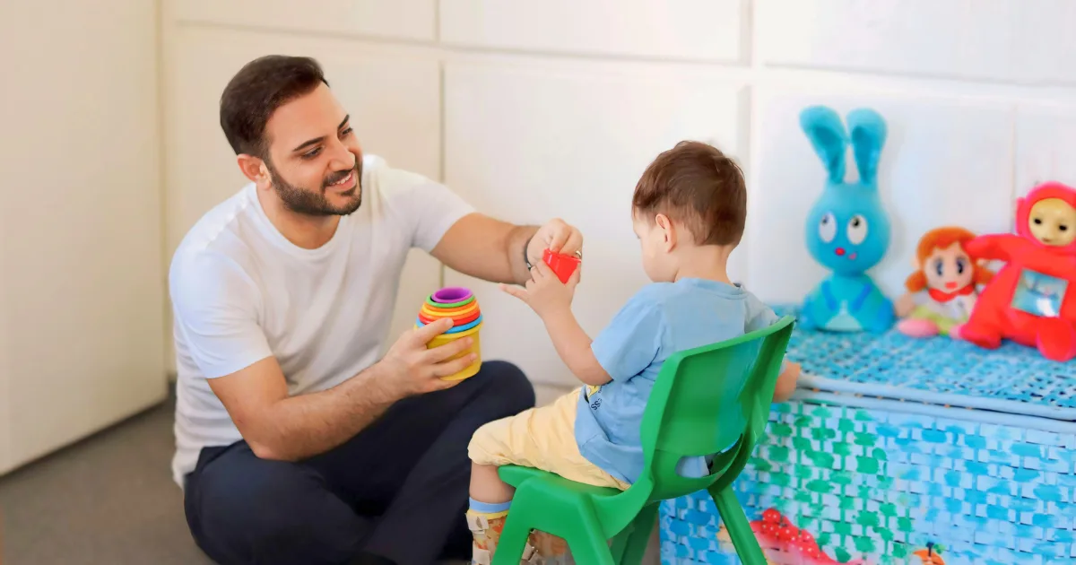 A man smiles while engaging fathers' role, playing with a young child seated on a green chair. They are stacking colorful rings, with plush toys and a blue storage basket in the background.