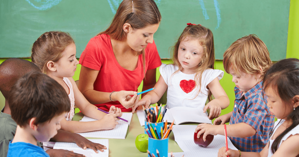 A teacher sits at a table with six young children, guiding them through a writing activity in a classroom. As they draw and write in notebooks, the group explores simple ethical dilemmas, with colored pencils, apples, and a green chalkboard nearby.