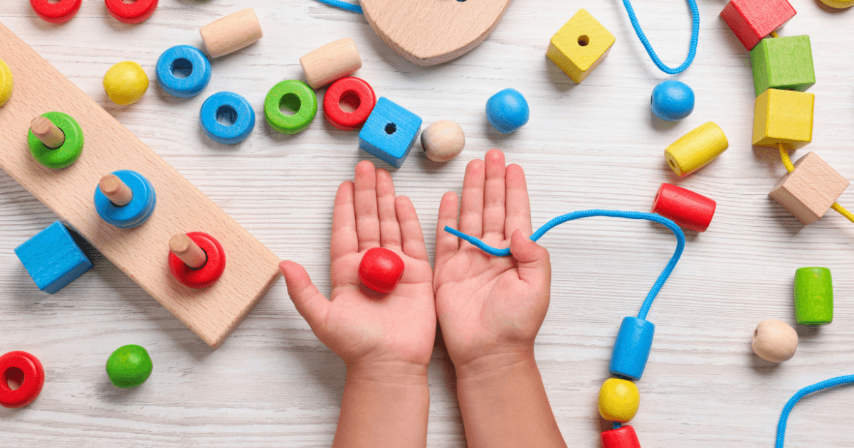 Child’s hands holding a red wooden bead and blue string, surrounded by colorful wooden beads and shapes on a white surface, with a lacing toy and stacking puzzle that help develop executive function skills.