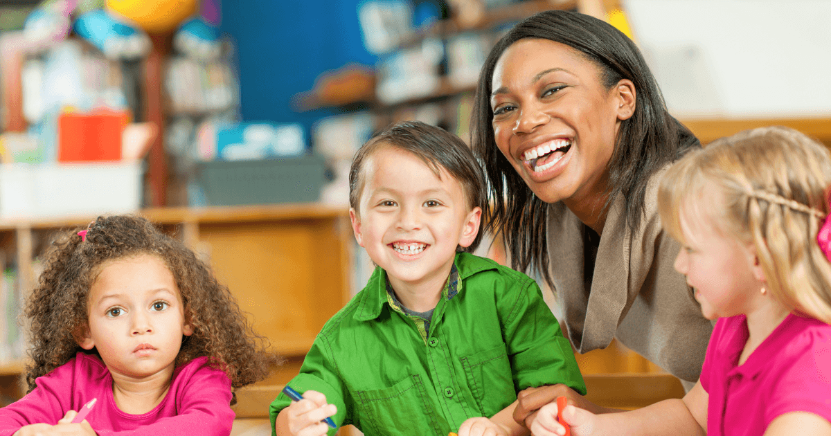 A smiling teacher sits at a table with three young children in a classroom at one of the faith-based preschools. Two children are smiling, while one looks serious. Art supplies fill the table, and bookshelves line the background.