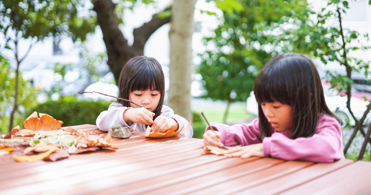 Two young children sit at a wooden table outdoors, drawing or writing on autumn leaves with pens—one of many engaging fall sensory activities. Fallen leaves are scattered on the tabletop, with green trees visible in the background.