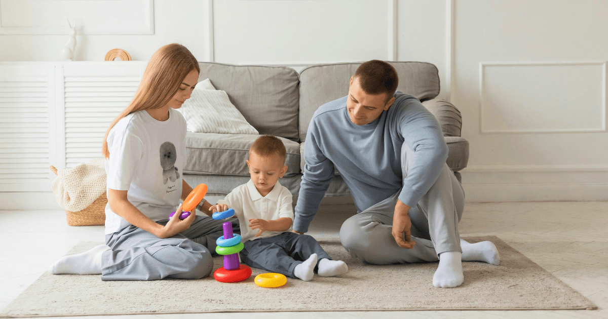 A woman, a man, and a young child sit on a rug in the living room, enjoying family engagement as they play with a colorful stacking ring toy. A gray couch and light-colored decor complete the cozy scene in the background.