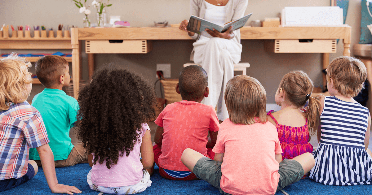 A group of young children sit on the floor in a classroom, facing a teacher who is reading a book to them during family literacy night. The teacher is seated in front, and the children are listening attentively.