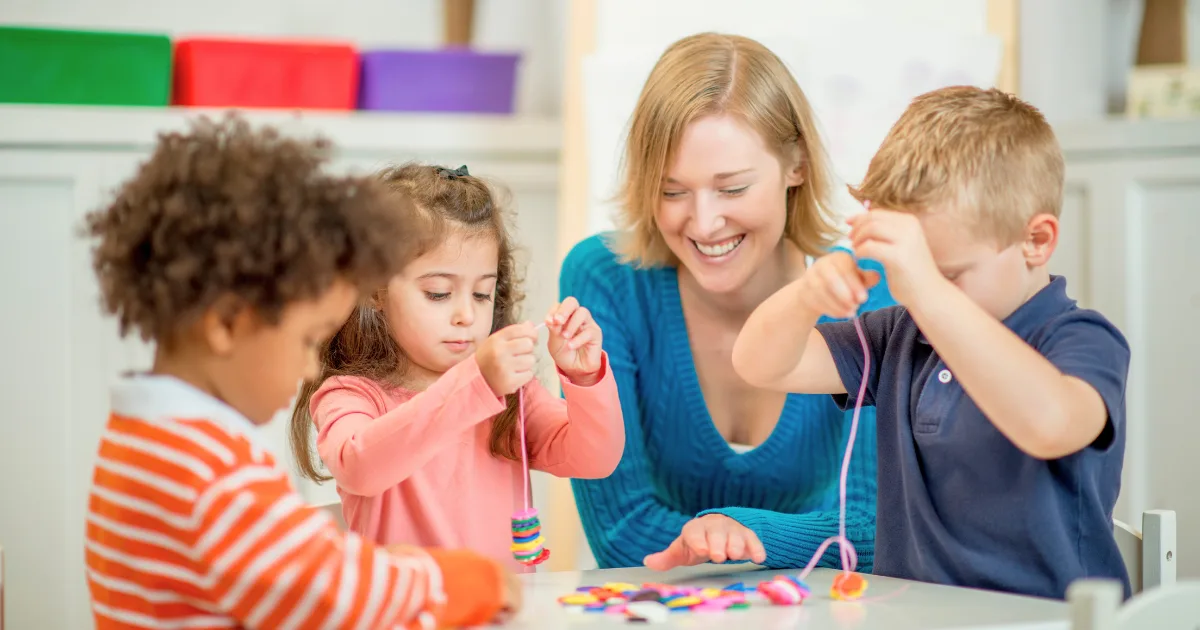 A woman smiles while sitting at a table with three young children, who are focused on stringing colorful beads together—one of many engaging fine motor activities for preschoolers.