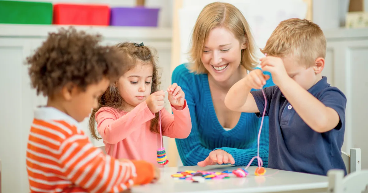 A woman smiles while watching three young children sitting at a table, threading colorful beads onto strings in a bright classroom setting to help develop their fine motor skills.
