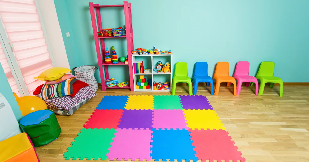 A bright, colorful playroom with flexible seating options like small colorful chairs, a foam mat of multicolored squares on a wooden floor, shelves with toys and books, and a cozy pile of pillows in the corner.