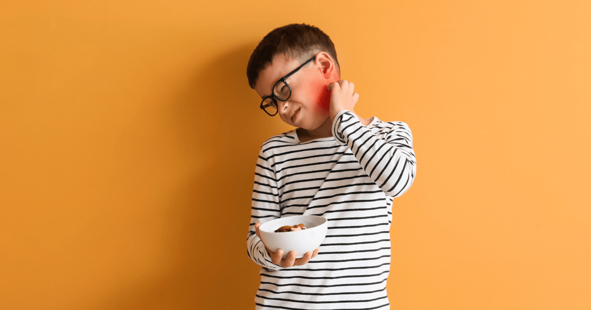 A boy wearing glasses and a striped shirt holds a bowl of food in one hand while scratching his red neck against an orange background. He looks uncomfortable, possibly experiencing food allergies.