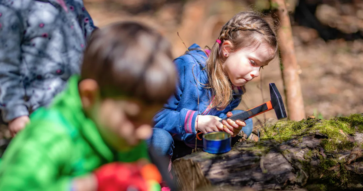 A young girl in a blue jacket uses a small hammer on a log at a forest preschool, while another child in green is blurred in the foreground. The scene takes place in a wooded area.