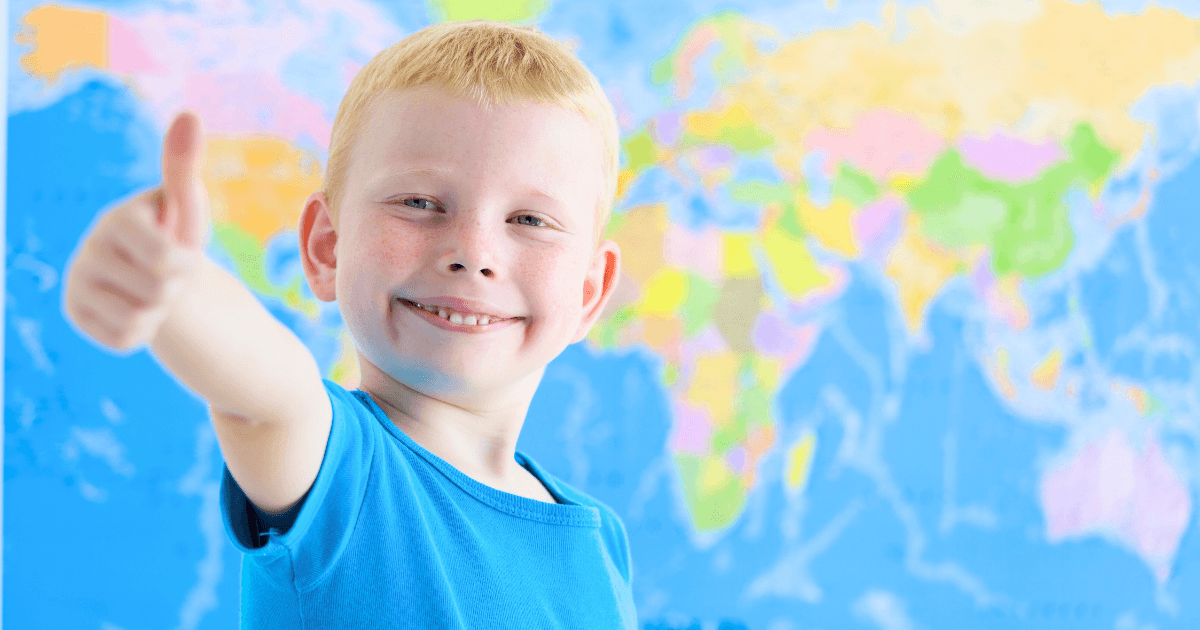 A smiling young boy with light hair wearing a blue shirt gives a thumbs up in front of a brightly colored world map, embracing the spirit of global citizenship.