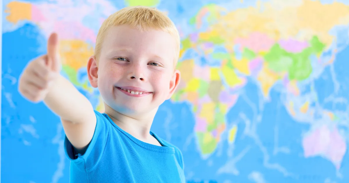 A smiling young boy with light hair wearing a blue shirt gives a thumbs up in front of a brightly colored world map, embracing the spirit of global citizenship.