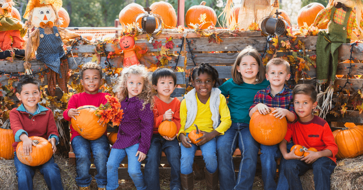 A group of seven smiling children sit on hay bales holding pumpkins, surrounded by autumn leaves, scarecrows, and festive harvest festival decorations.