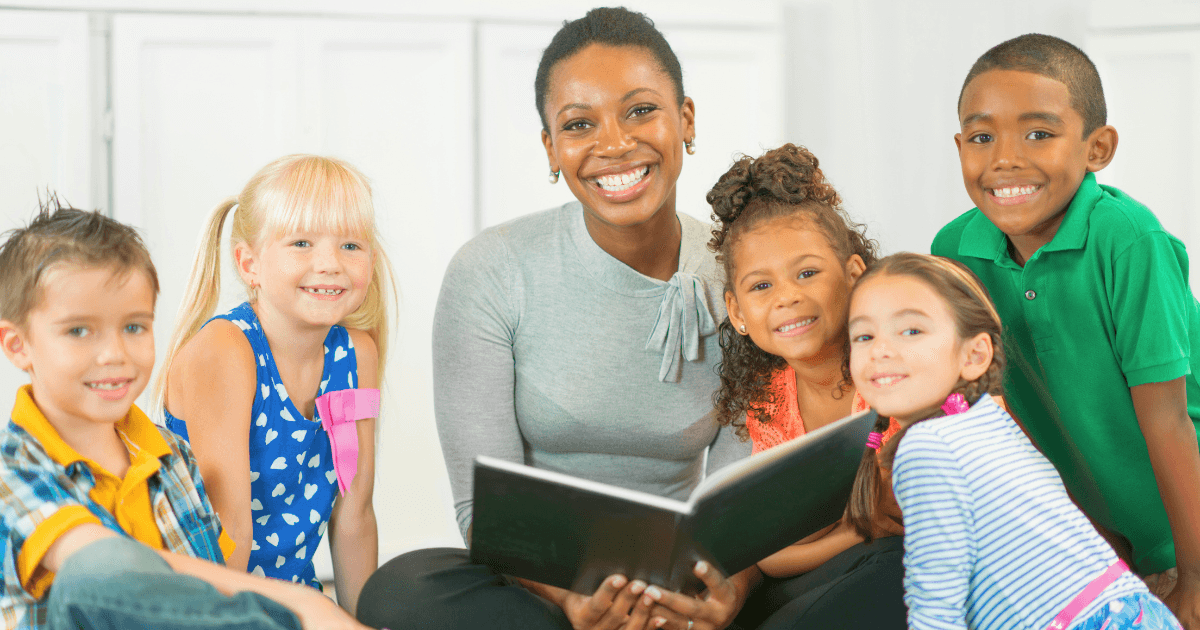A smiling woman sits on the floor of a high-quality preschool, holding an open book and surrounded by five happy children of diverse backgrounds in casual clothing, suggesting a storytime or reading activity together indoors.