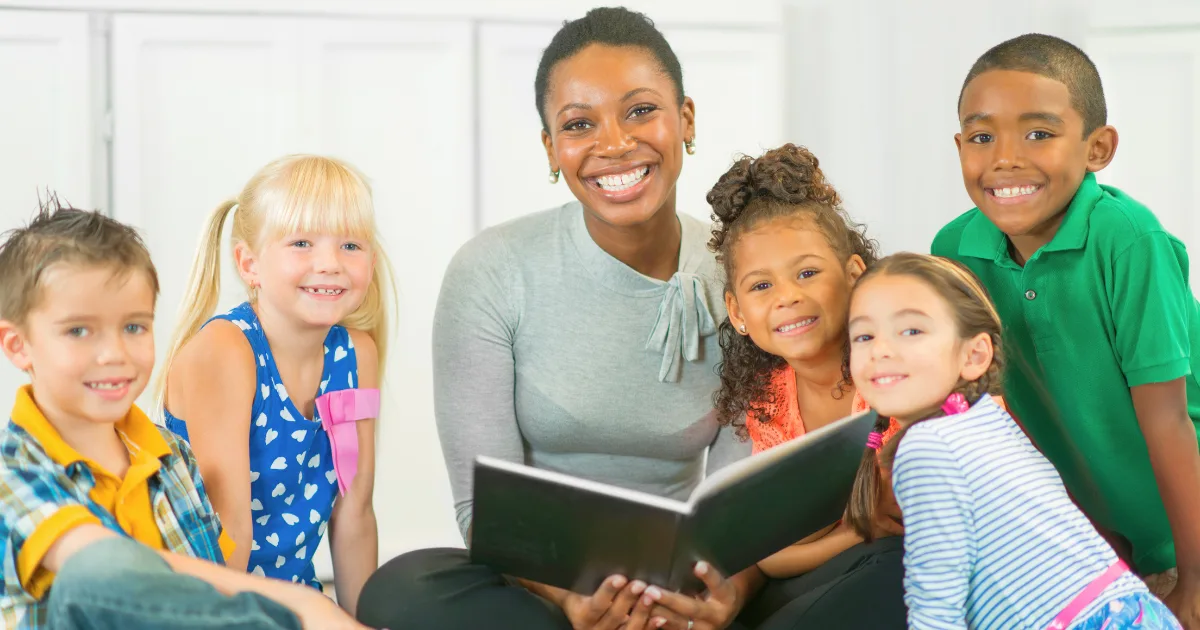 A smiling woman sits on the floor of a high-quality preschool, holding an open book and surrounded by five happy children of diverse backgrounds in casual clothing, suggesting a storytime or reading activity together indoors.