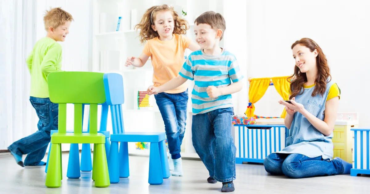 Three young children play musical chairs indoors at a quality preschool, smiling and moving around colorful chairs, while a woman nearby claps and encourages them. The bright room is filled with toys and books.