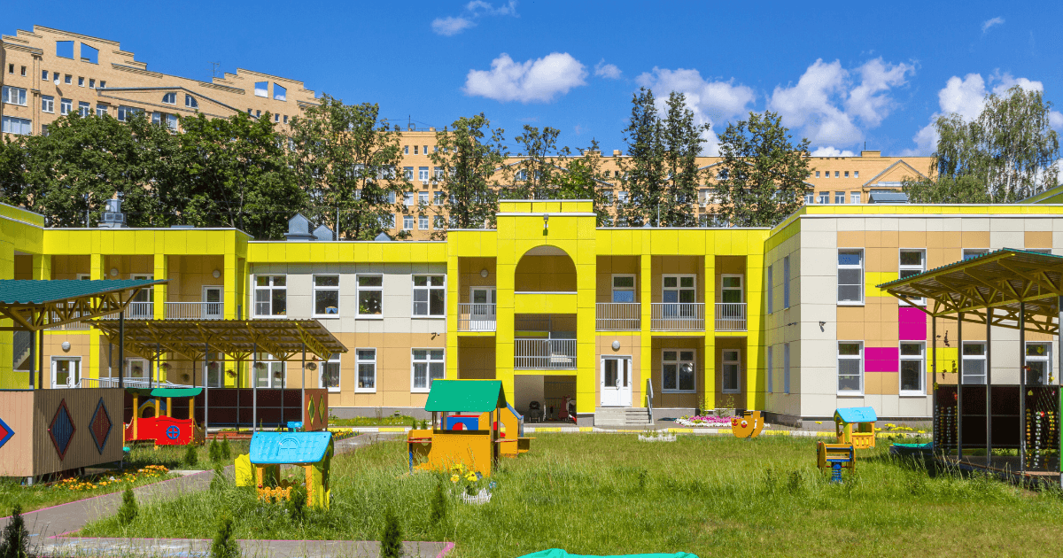 A modern, yellow and beige building with playground equipment in front, surrounded by grass and trees—a picture-perfect setting for parents learning how to choose a preschool, under a sunny blue sky with scattered clouds.