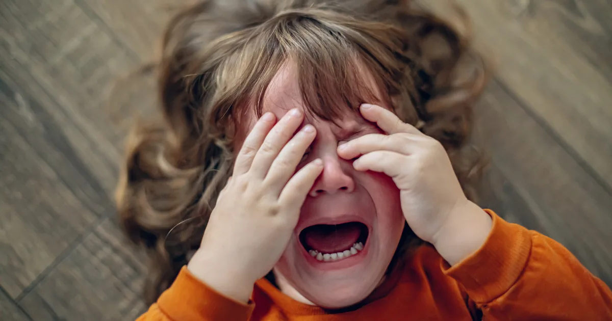 A young child with long brown hair lies on a wooden floor, crying with their mouth open and hands covering their eyes, wearing an orange long-sleeve shirt—an all-too-familiar scene of preschool tantrums.