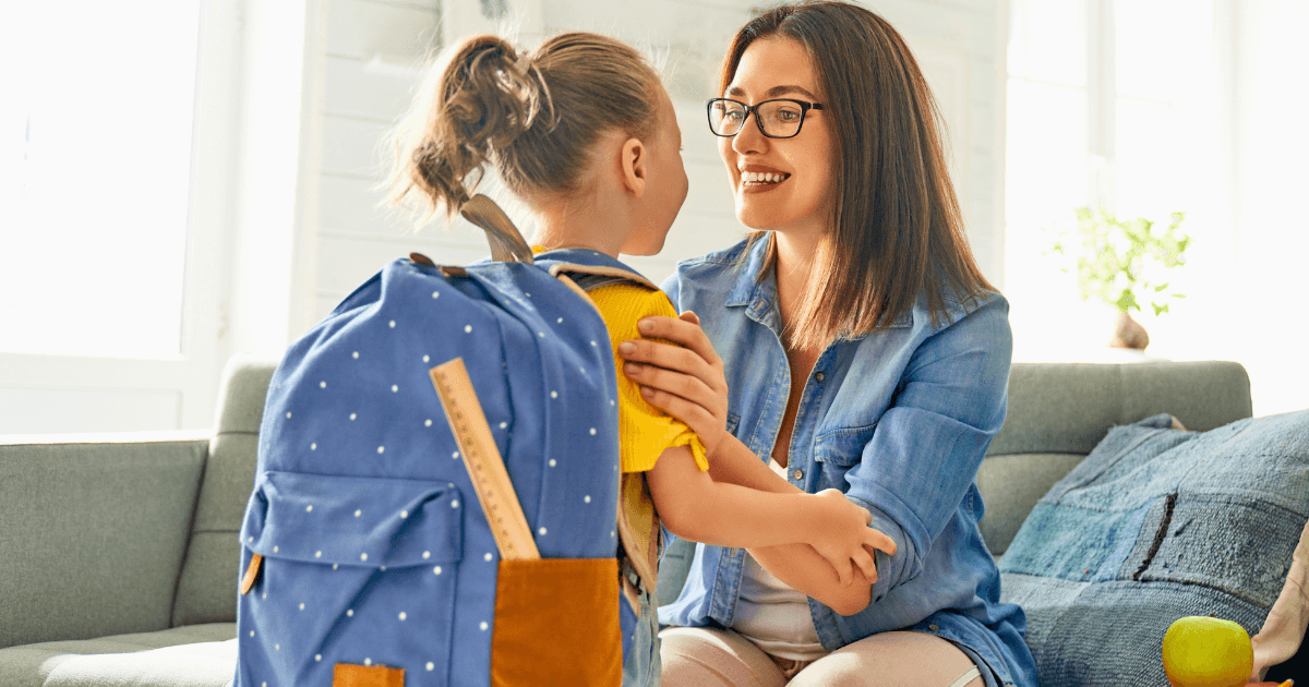 A woman, smiling, kneels in front of a young girl with a blue backpack, holding her shoulders. They are indoors on a couch, sharing a happy moment—showing how to prepare your child for preschool with warmth and encouragement.
