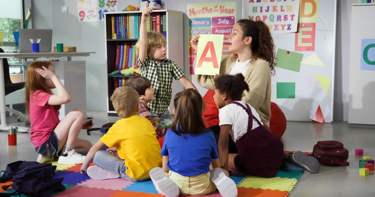 A teacher sits on the floor with a group of young children, demonstrating how to teach letters to preschoolers by holding up a card with a large red letter A while the attentive class listens. Educational posters and toys brighten the background.