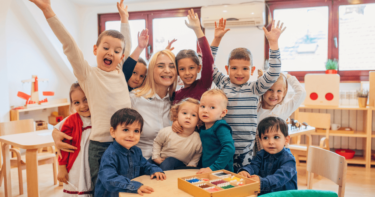 A group of young children and a smiling adult sit and stand together in a bright, inclusive classroom, with some children raising their hands excitedly. Educational toys and chairs are visible in the background.