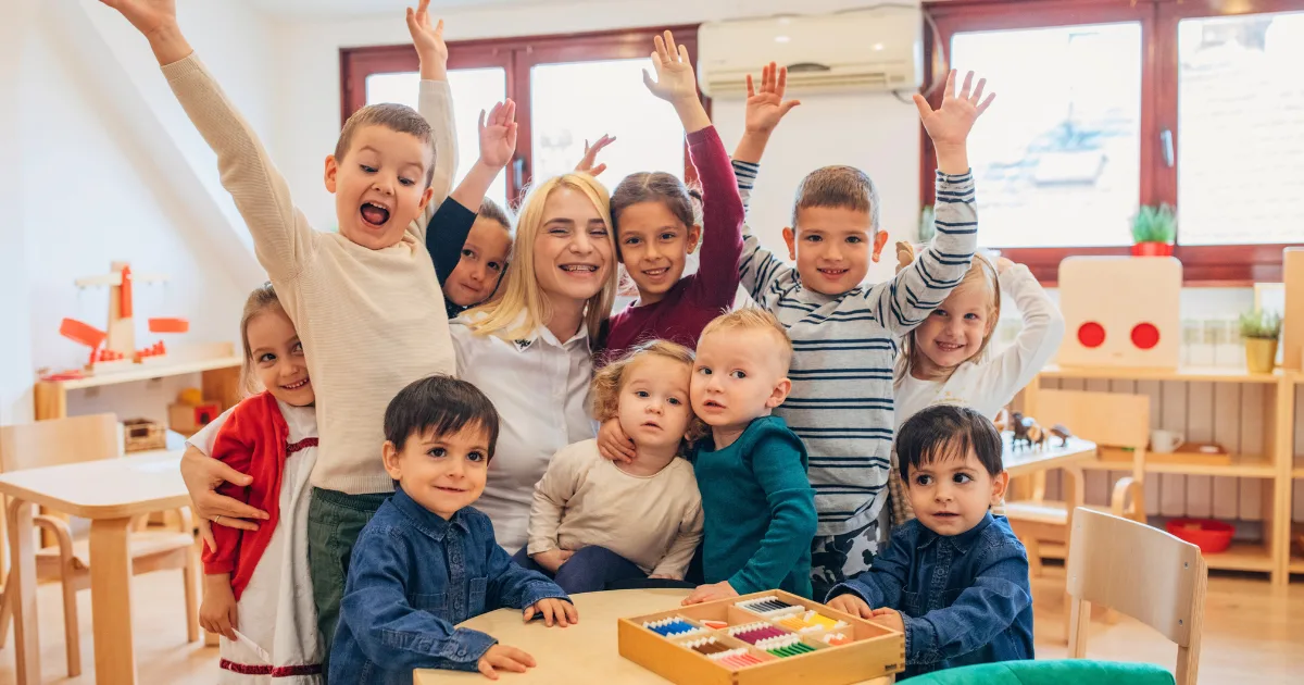 A group of young children and a smiling adult sit and stand together in a bright, inclusive classroom, with some children raising their hands excitedly. Educational toys and chairs are visible in the background.