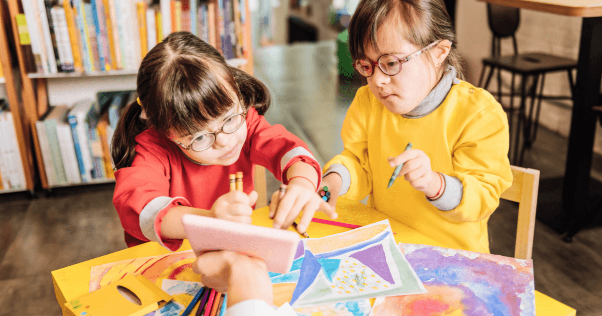Two young girls wearing glasses and colorful clothes sit at a table in an inclusive preschool library, drawing and coloring with pencils and crayons on paper, surrounded by books and art supplies.