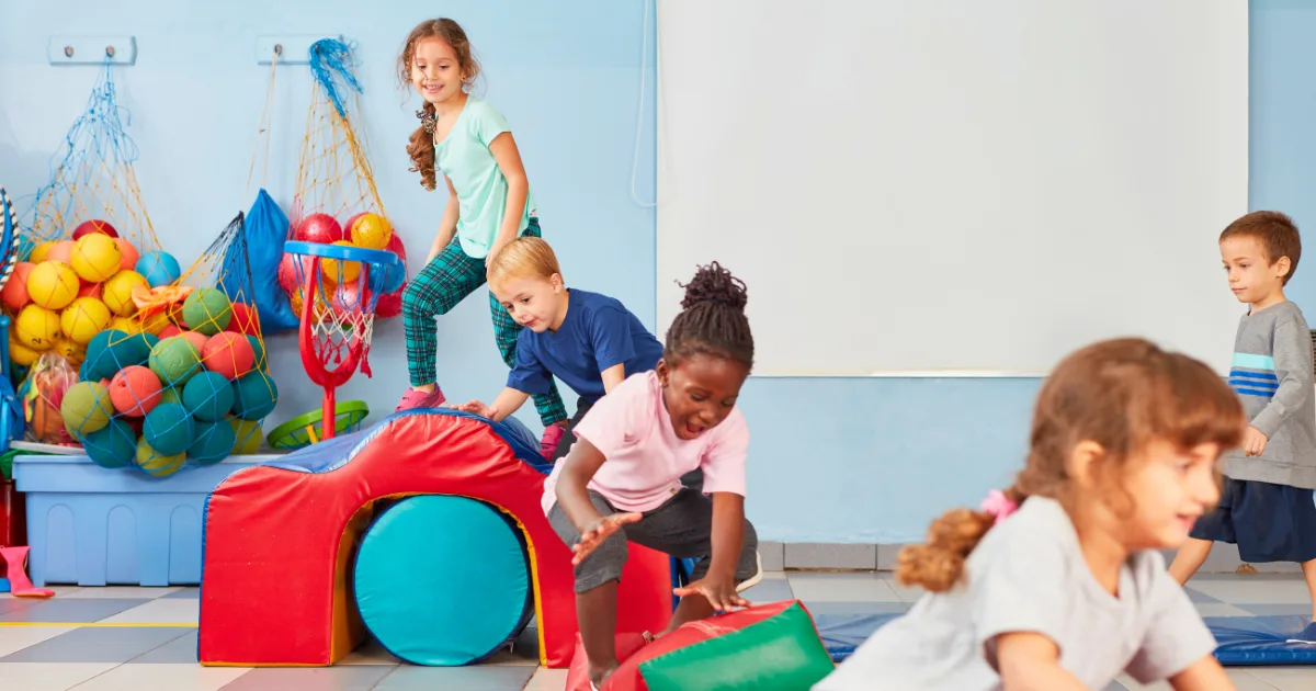 Children play and climb on colorful soft play equipment in a bright indoor playroom, enjoying indoor movement activities. A large bin of balls and nets is visible in the background as they appear energetic and joyful.