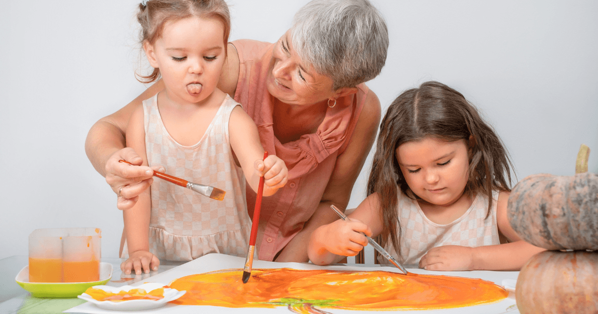 An older woman smiles while helping two young girls paint a large orange pumpkin shape on paper as part of intergenerational programs. One girl sticks out her tongue while painting, and the other focuses on her brushwork. A pumpkin sits on the table.