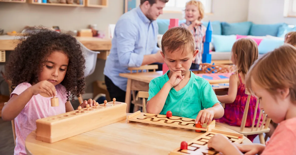 Children sit at a table in a classroom, concentrating on wooden educational puzzles, reflecting environments where parents wonder, "Is Montessori worth it?" The cozy, colorful room is filled with children and an adult engaging in activities.