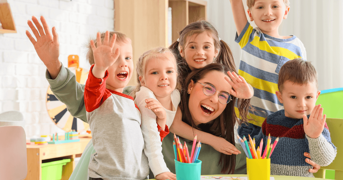 A group of happy children and a smiling adult sit at a table with colored pencils, waving at the camera in a bright, cheerful classroom focused on kindergarten readiness.