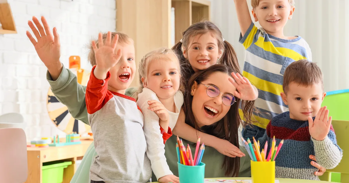 A group of happy children and a smiling adult sit at a table with colored pencils, waving at the camera in a bright, cheerful classroom focused on kindergarten readiness.