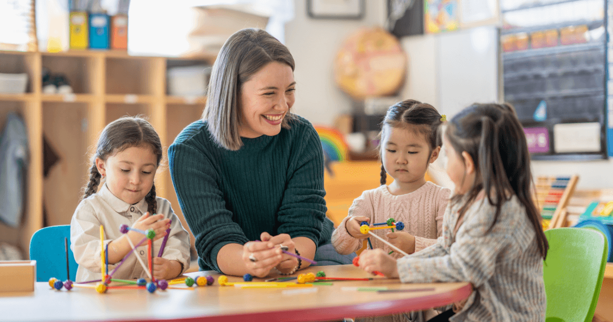 A teacher sits at a table with three young children during a kindergarten visit, smiling and engaging with them as they build structures with colorful sticks and balls. Shelves with educational materials are in the background.