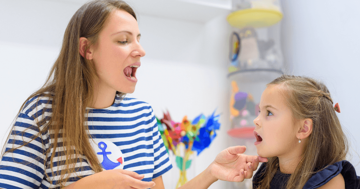 A woman and a young girl sit facing each other, both with their mouths open as if practicing speech sounds. The woman gently holds the girl’s chin, guiding her in a language development exercise in a bright room.