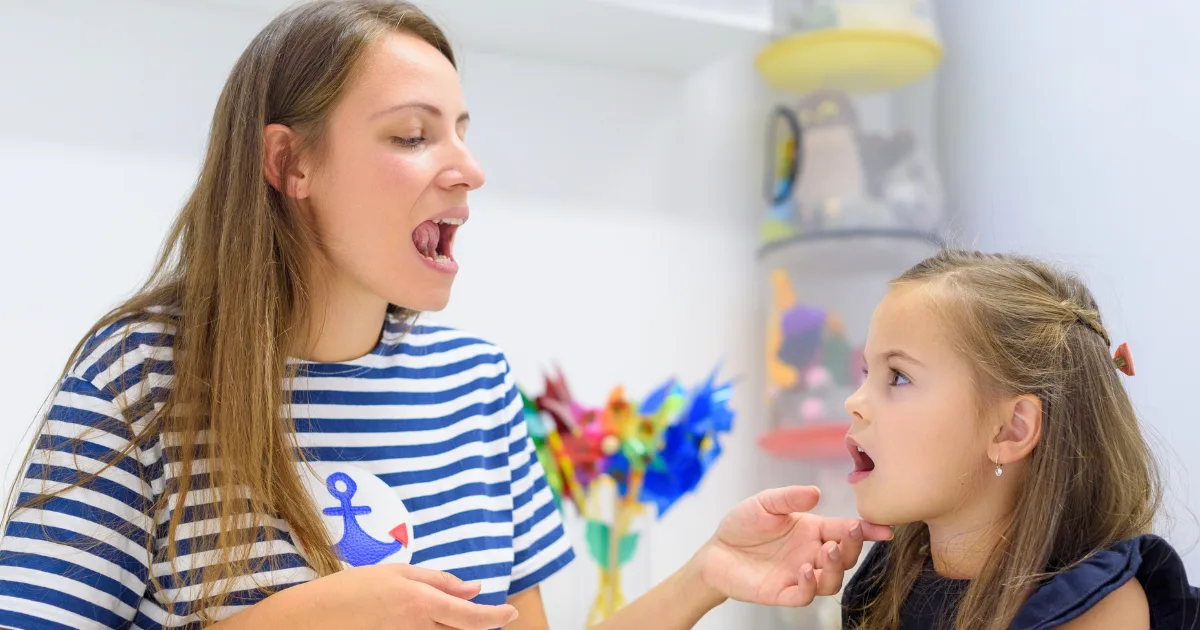 A woman and a young girl sit facing each other, both with their mouths open as if practicing speech sounds. The woman gently holds the girl’s chin, guiding her in a language development exercise in a bright room.