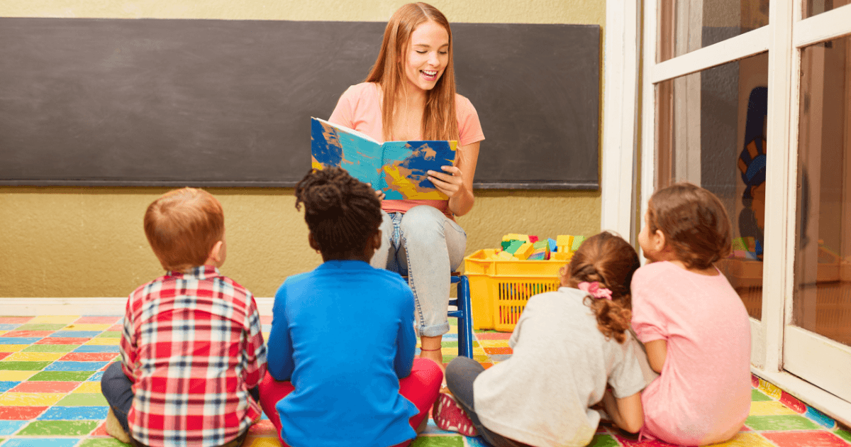 A young woman sits on a chair reading learning stories from a colorful book to four children seated on a vibrant tiled floor in a classroom, with a blackboard and large window in the background.