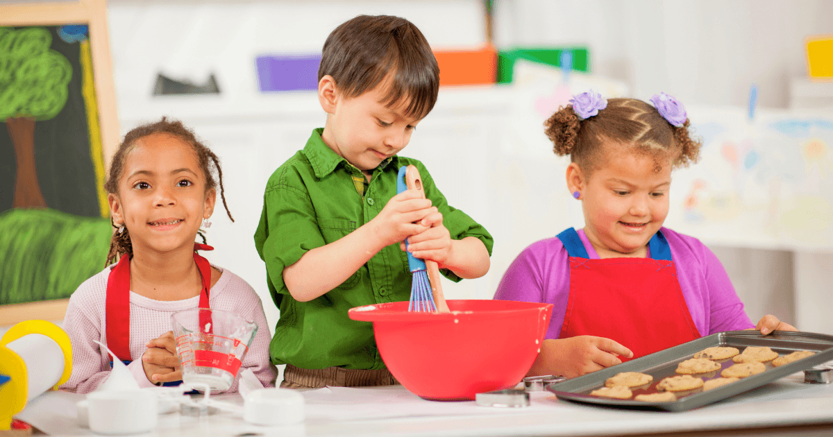 Three young children engage in literacy-rich dramatic play as they bake together in a kitchen. One whisks in a red bowl, another holds a tray of cookies, and the third measures ingredients—all smiling and wearing aprons.
